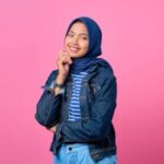 A cheerful young woman wearing hijab and denim jacket against a pink background, smiling warmly.