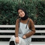 Young Asian woman in hijab sitting on a bench, smiling against lush greenery, exuding a peaceful outdoor vibe.